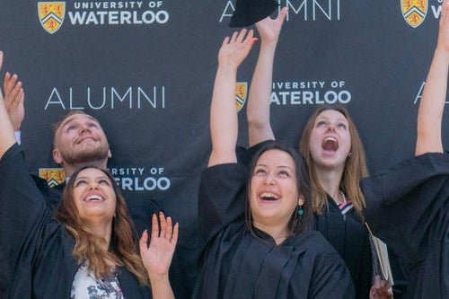 Graduates throwing their caps in the air in front of an University of Waterloo Alumni setp and repeat