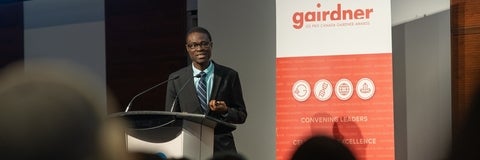 Link, Dr. Abiola Olaitan speaking in front of a crowd. There is a pull up banner behind him that says "gairdner" in larger letters and "LES PRIX CANADA GAIRDNER AWARDS" in smaller letters. 