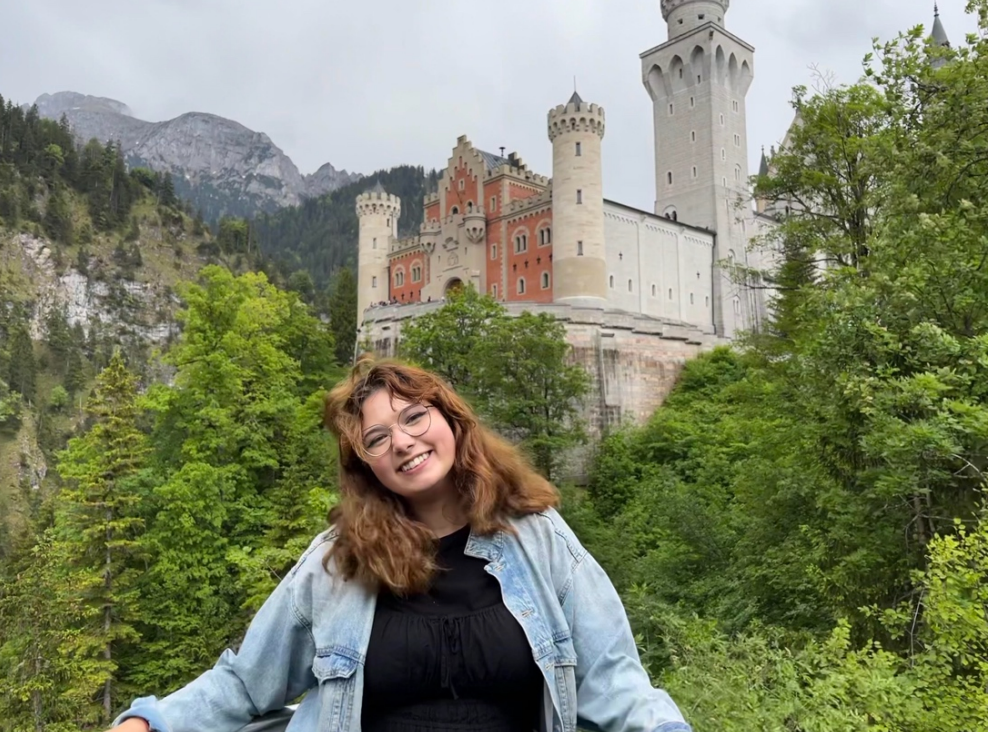 Person standing outside with a castle and mountains in the background. 
