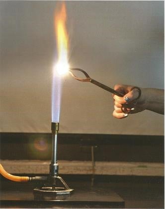 Tongs containing magnesium held over Bunsen flame by outstretched hand.