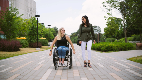 A woman walks next to another woman in a wheelchair on a walkway.