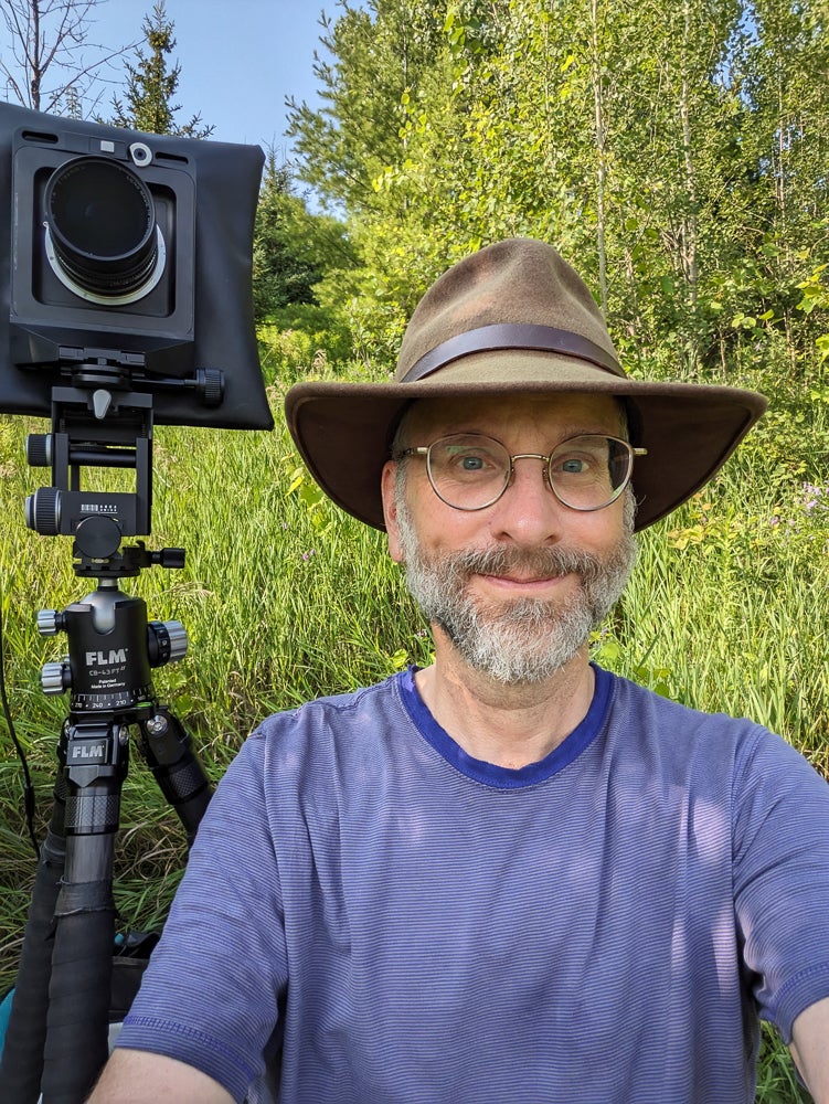 Rob de Loe standing next to a camera on a high tripod with forest and grasses in the background.