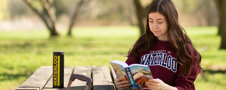 A student wearing a maroon sweater reads a travel guide at a table outside