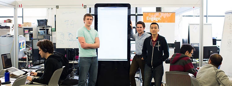 Business students stand in front of large display screen in cluttered business incubator