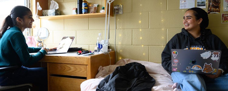 Two students sitting in a residence room. The wall behind one of the students is deocorated with photos and hanging string lights.