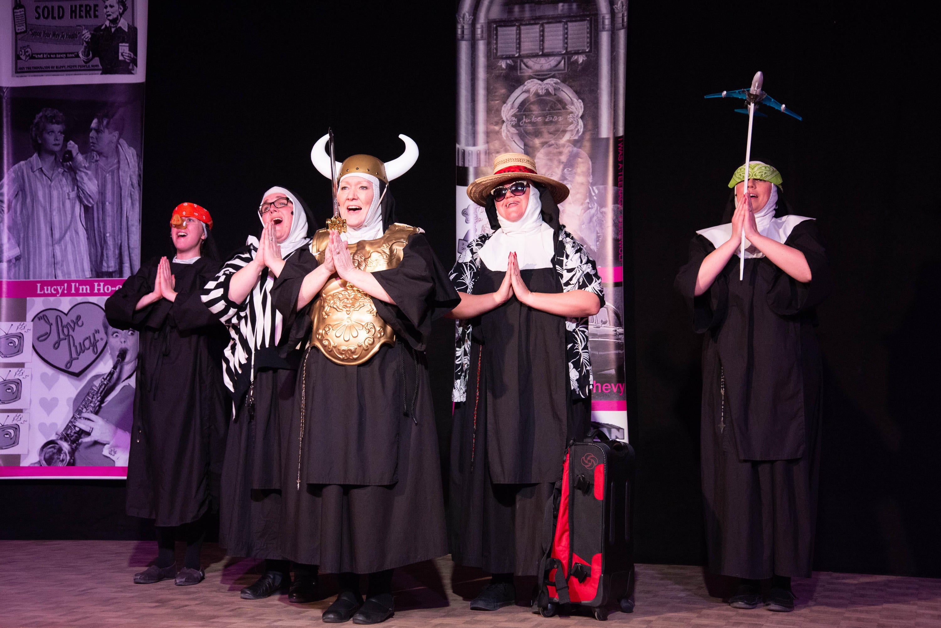 A group of actresses dressed as nuns sing on stage with props like viking horns, a model airplane, and traveling gear.