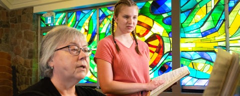 A student listen's to their singing instructor in the chapel. Stainglass windows ligh up the background.