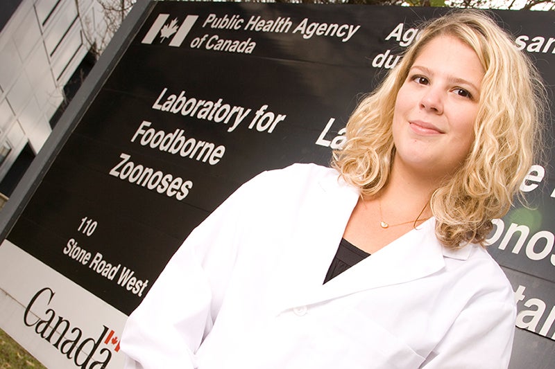 Co-op student in lab coat stand in front of Public Health Agency of Canada lab sign.