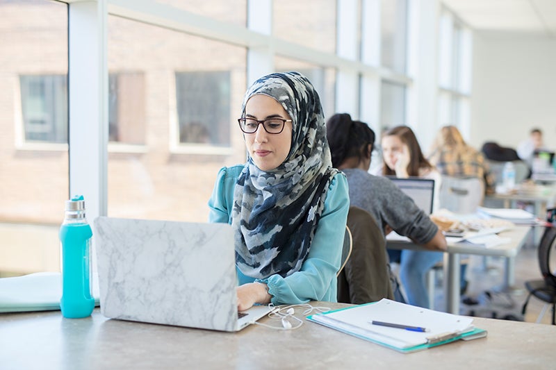 Student wearing hijab works on lab top with other students in distance.