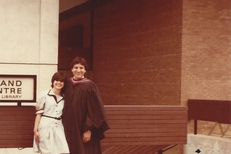 Man in graduation gown stands with woman in front of building.