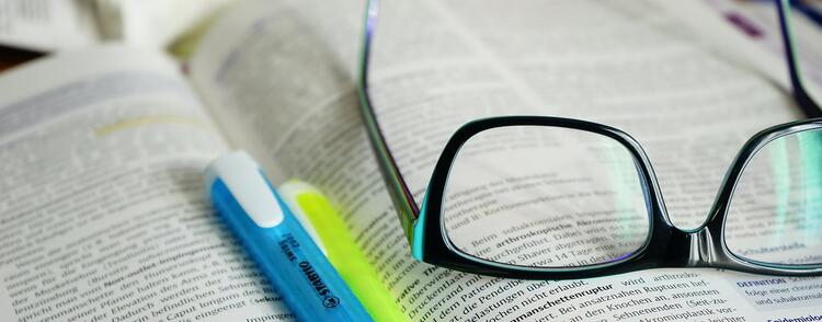 An open textbook, with a pair of glasses laying upside on the right hand side and a few pens lying in the centre. 