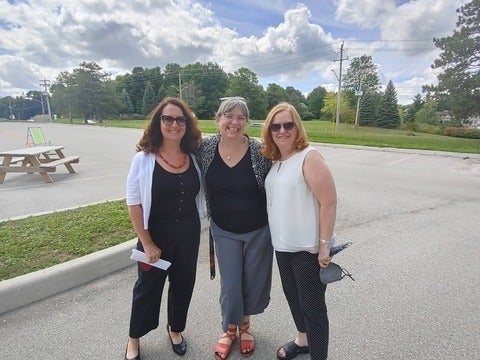 Three women standing in a group in the Renison parking lot on a sunny day, smiling at the camera. 