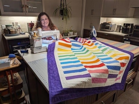 A woman sits at a table with a sewing machine. A large rainbow quilt is spread out on the able in front of her.