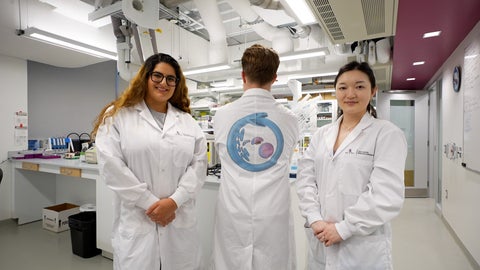 Three scientists in the NeurdyPhagy lab. A male scientists is standing with his back turned. He is wearing a white lab coat with a design on the back. Two female scientists are standing on either side facing the camera. They are wearing white lab coats.  