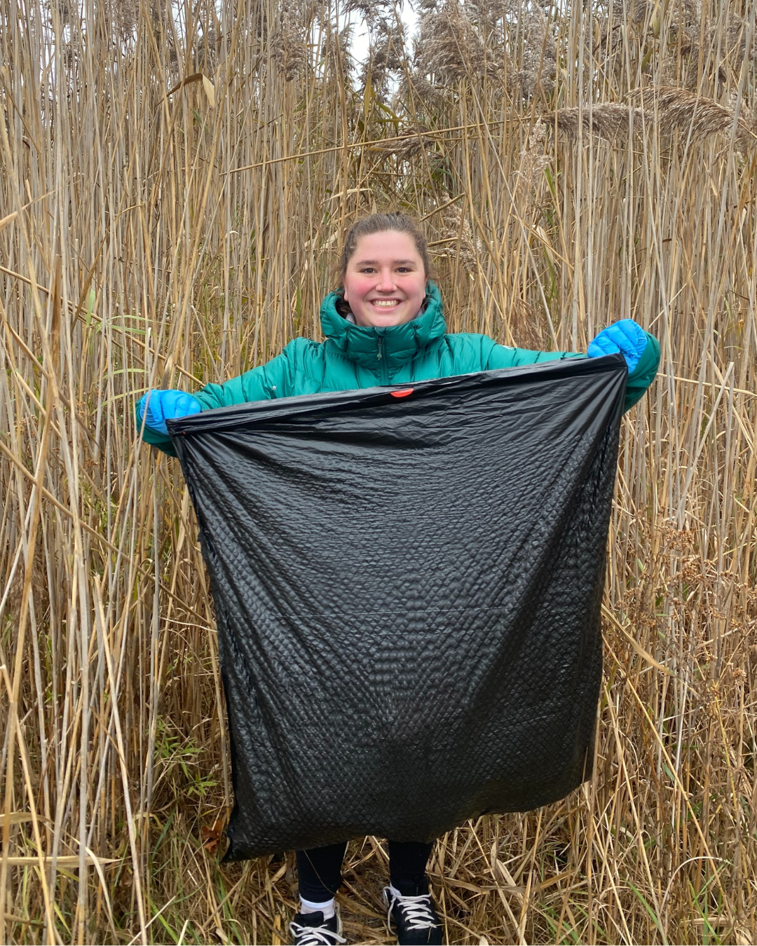 Woman in teal coat holding up a trash bag in a wetland.