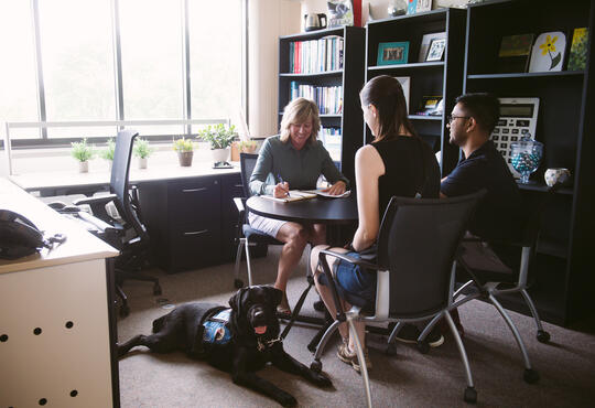 Two students with guide dog talking to woman at a desk