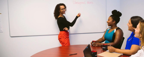 A woman at a whiteboard surrounded by three women