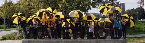 Smiling staff holding black and gold umbrellas stand behind Waterloo sign