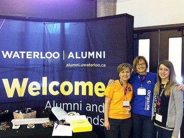 Three female alumni standing in front of Waterloo Welcome banner