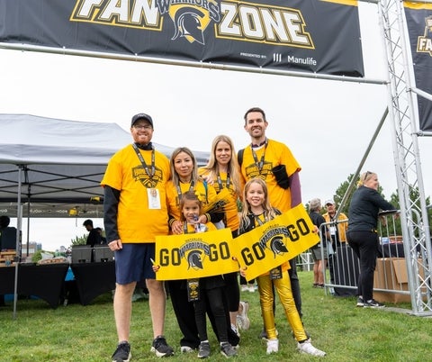 A family poses in front of the Fan Zone, all wearing Warriors gear and holding Warriors signs