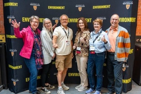 Six alumni smile in front of an Environment photo wall