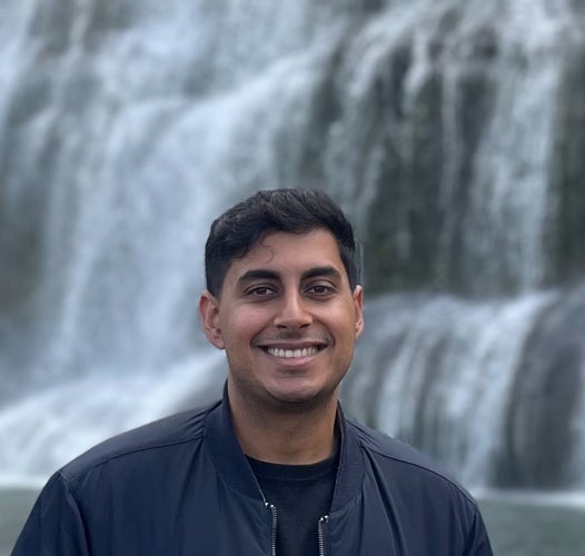 Young man with black hair, and medium to dark skin smiling, standing in front of a waterfall wearing a black tshirt and navy blue jacket
