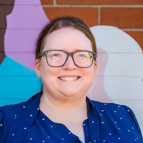 Image of Katie (Waterloo Chapter Member) standing in front a colorful brick wall wearing a blue and white polka dot shirt