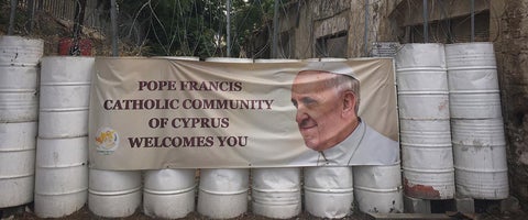 A row of white barrels with a sign on it that has a welcome from the Pope's Catholic Community in Cyprus