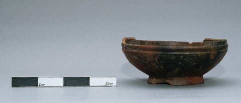 A small clay bowl artifact next to a black and white ruler to show what size it is