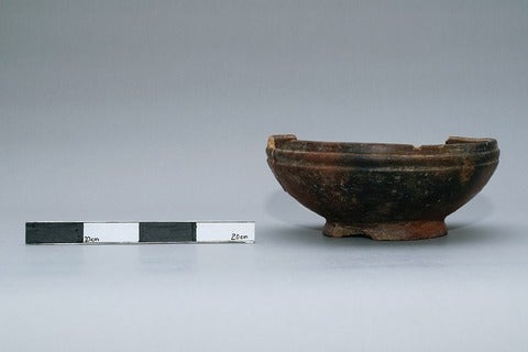 An artifact (a brown clay bowl) sits next to a black and white ruler to show what size it is
