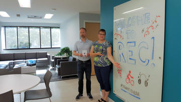 Troy and Sandy in front of 2 large whiteboards, dining tables and lounge.