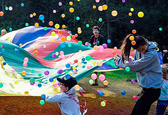 Camp staff and kids bounce balls on a parachute.