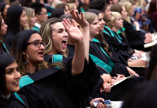 Graduates at convocation wearing robes and smiling and waving.