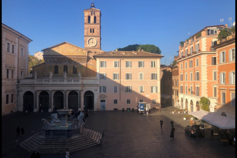 A historic Italian square with a central stone fountain. In the background is a church with arched entrances and a tall brick bell tower with a clock. Surrounding the square are multi‑story pastel buildings. A few people walk across the open cobblestone plaza, and a small market stall is set up on the right.