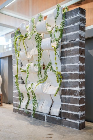 A vertical brick-framed panel holds a grid of white, interlocking ceramic modules. Several strands of trailing green plants weave through the openings, hanging down the front of the structure. The piece is displayed indoors on a concrete floor, with shelves and other objects faintly visible in the background.