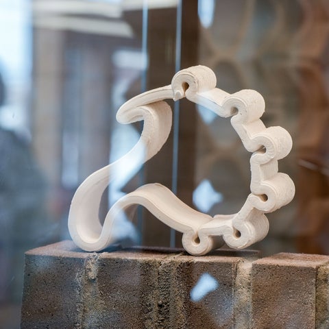 A small white ceramic sculpture with looping, interlocking curves sits on a textured brick base. The form twists in a continuous ribbon-like shape, creating rounded openings along its edge. The piece is displayed inside a glass case with a softly blurred background.