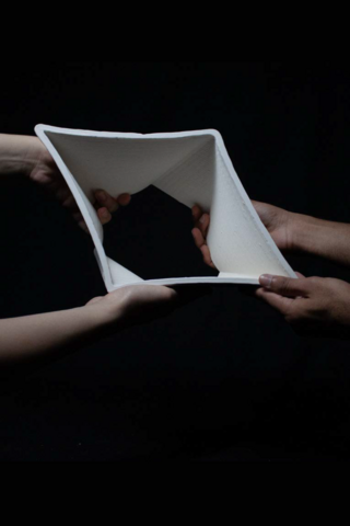 Two pairs of hands hold the corners of a hollow, square-shaped ceramic form against a dark background. The object is slightly twisted, creating an angled opening in the center as the hands support it from each side.