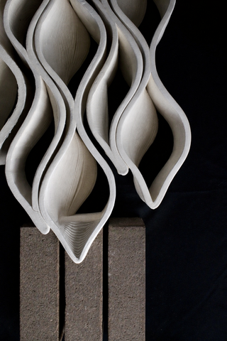 A close-up view of a sculptural wall screen made of repeating wavy, leaf‑shaped ceramic modules, with warm light shining through the openings. The screen sits beside a dark grey brick surface.