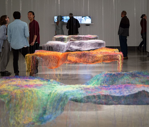 interior of the Riverside Gallery at the school of architecture, four multicoloured fabric art pieces are hung in a row in the centre of the photo while seven people look around the gallery