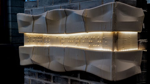 A horizontal ceramic light fixture mounted on a brick wall. The fixture consists of textured ceramic panels framing a central illuminated strip with small raised bumps along its surface.