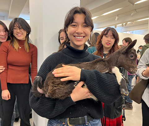 a smiling student holds a baby goat while several of their classmates watch