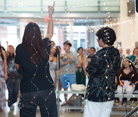 rear view of two students in front of an audience in the first floor atrium of the school of architecture, there are string lights in the foreground