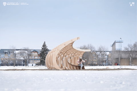 A wooden wave‑shaped installation made of curved, rib-like plywood frames sits on a snowy beach. A person is seated inside the structure, which opens toward the viewer