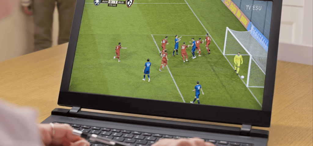 A person watches a soccer match on a laptop screen showing players in red and blue uniforms around the goal.