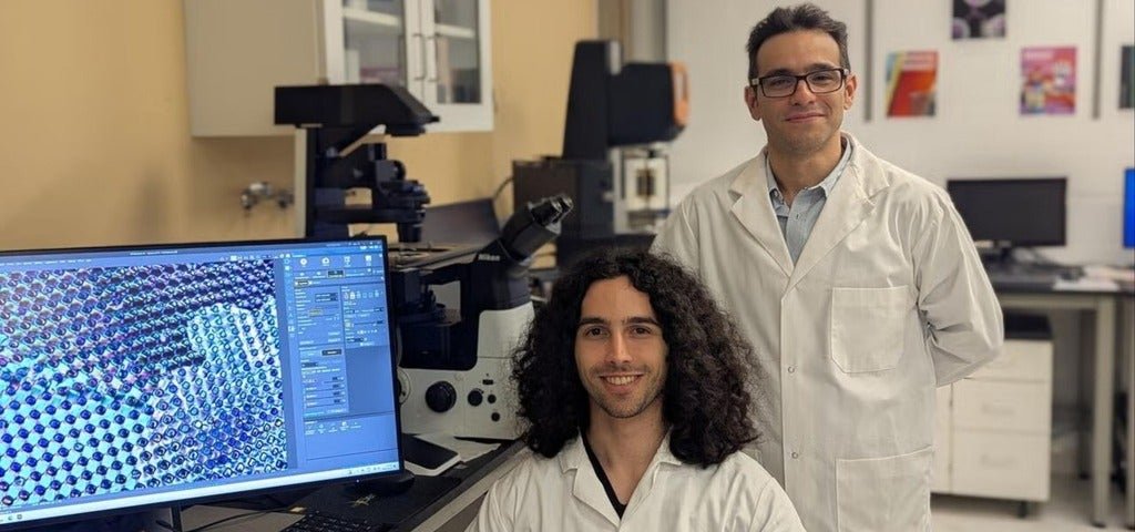 Two individuals, one standing and one seated, are in a laboratory setting wearing white lab coats. The person sitting in front of a computer is smiling, and a detailed microscopy image is displayed on the screen. Laboratory equipment is visible in the background.