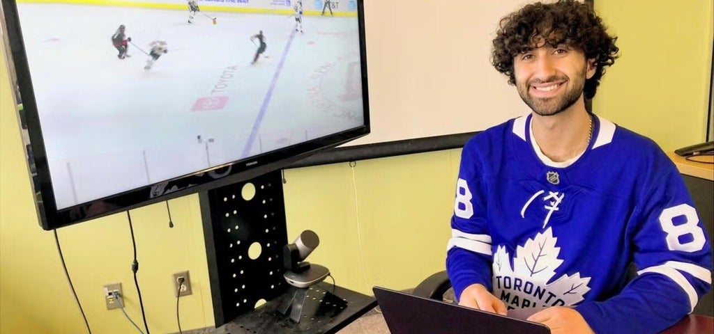 A person wearing a blue Toronto Maple Leafs jersey sits at a desk with a laptop, smiling at the camera while watching a hockey game on a large screen. The TV screen shows players in action on the ice.