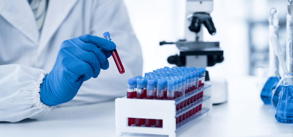 A gloved researcher holds a test tube filled with red liquid above a rack of similar tubes next to a microscope