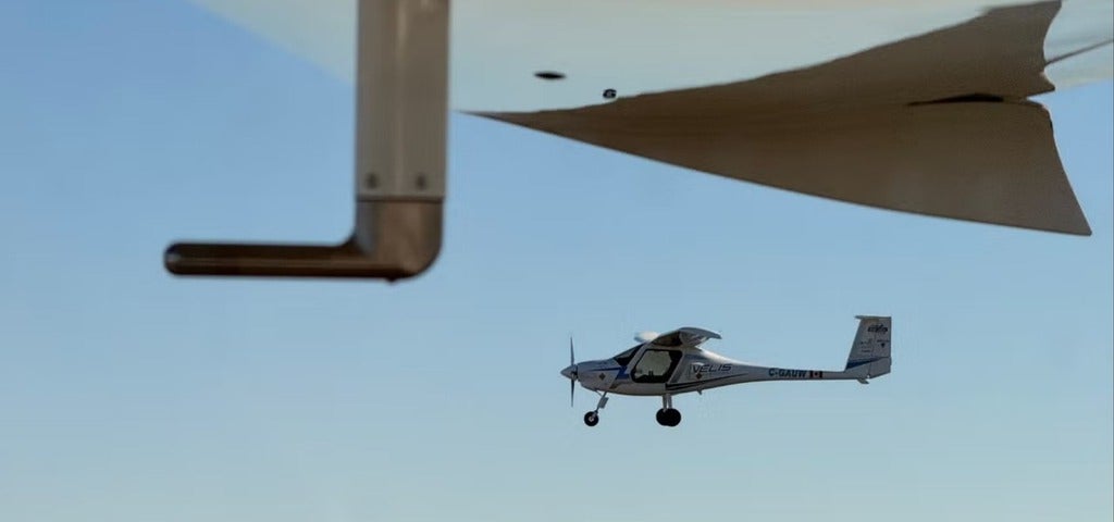 A small aircraft is flying in the sky, captured from a perspective looking at the underside of the airplane wing. The plane is identified as a Velis Electro, with a clear sky in the background.