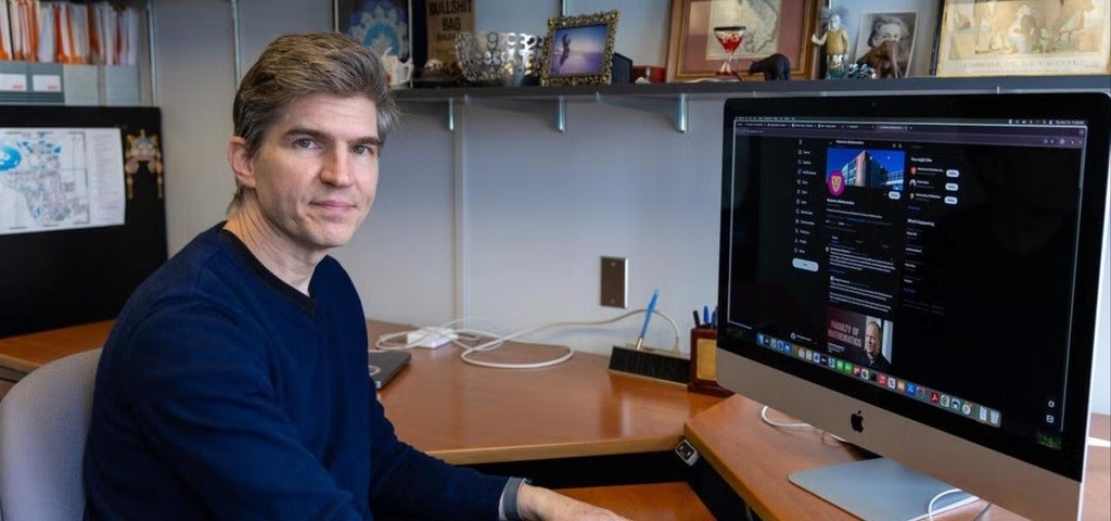 A person sits at a desk in front of an iMac, working on a computer. The desk is neatly organized with a few personal items and documents visible in the background. The individual appears focused on the computer screen.