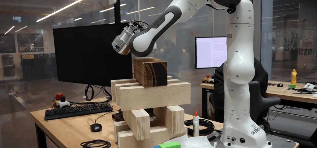 A robotic arm in a lab setting is positioned over a wooden block structure on a workbench, with computer monitors, keyboards, and lab equipment visible in the background.